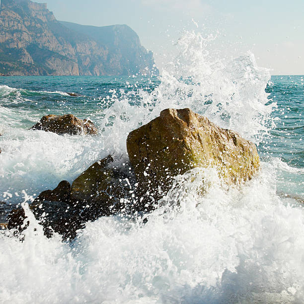 Waves crashing against rocks with sea foam and distant mountains.